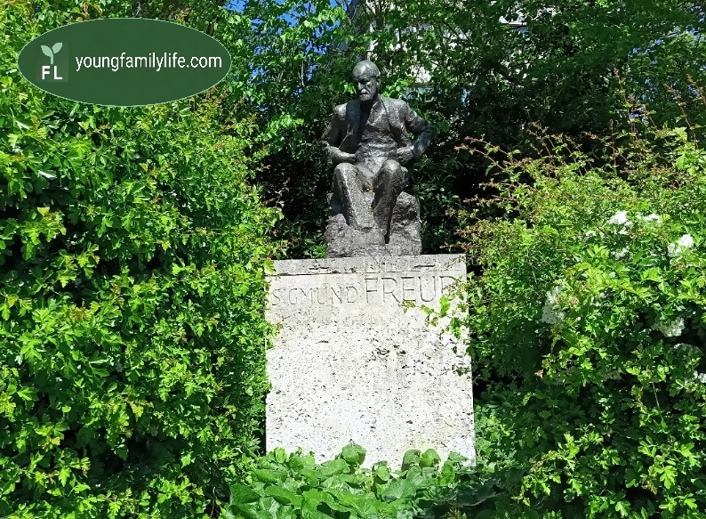 Bronze statue of Sigmund Freud, seated and contemplative, half-surrounded by dense green shrubbery outside the Tavistock Centre, London. Inscribed on the stone plinth: Sigmund Freud.
